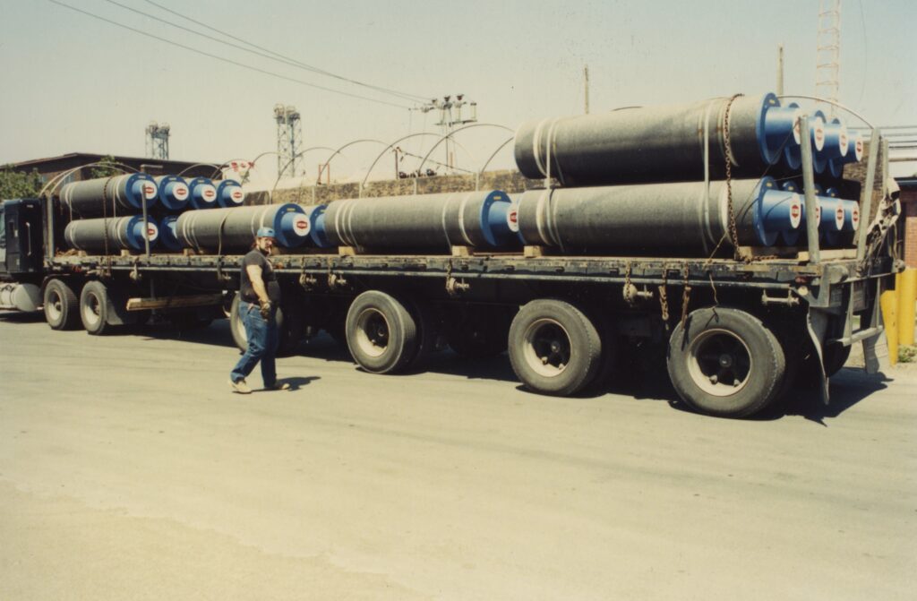 A man walks in front of a semi truck, carrying what looks like pipes.