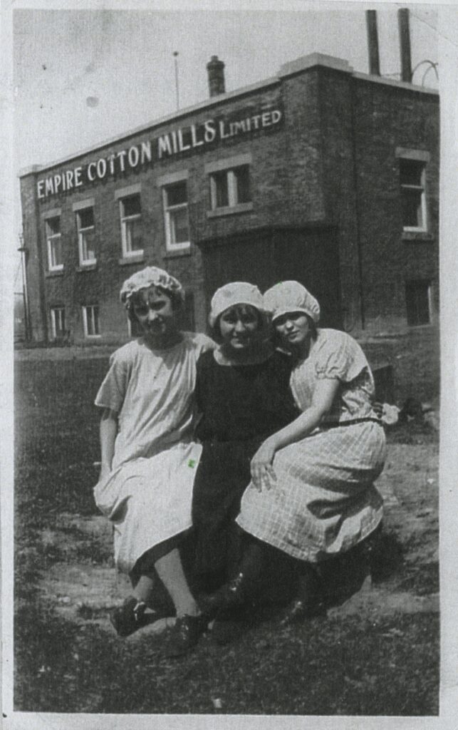 Three women in old fashioned dresses and bonnets pose together in front of a building which says "empire cotton mills limited". 