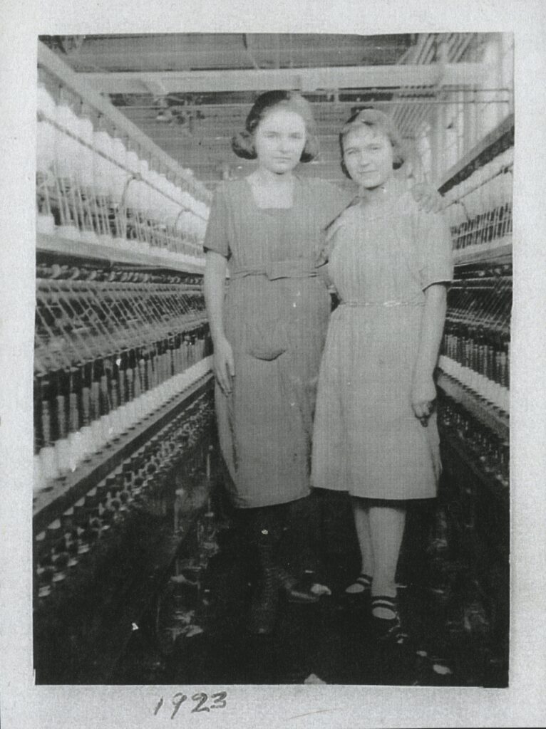 Two women in dresses pose with their arms around each other. They are standing between two rows of machinery in Empire Cotton Mills.