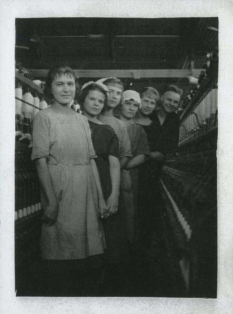 A group of women and one man pose together between two rows of machinery inside Empire Cotton Mills.