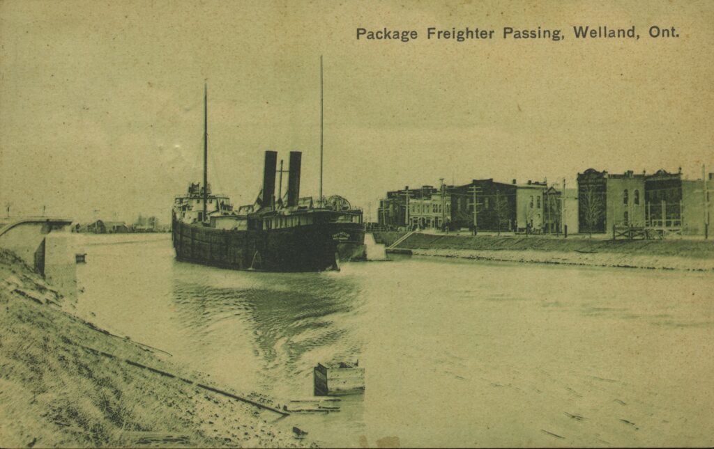 An old photo of a ship passing by houses on the canal. Text in the corner reads Package Freighter Passing, Welland.
