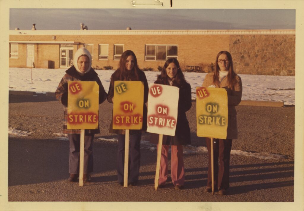 An old colour photograph of four women holding signs reading "UE on strike". There is snow on the ground behind them and they are dressed warmly.