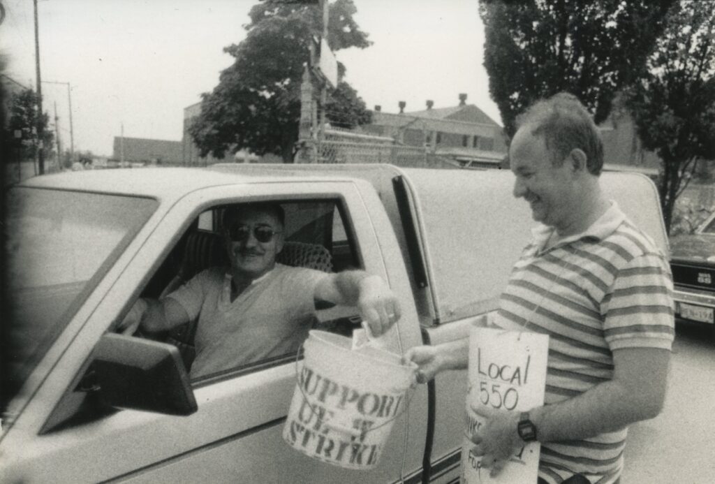 Two men at a strike. One can be seen through the window of his vehicle, the other is approaching the window. They hold between them a bucket.