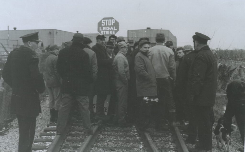 A group of men are gathered on railroad tracks. There is a sign behind them with a message relating to the strike.