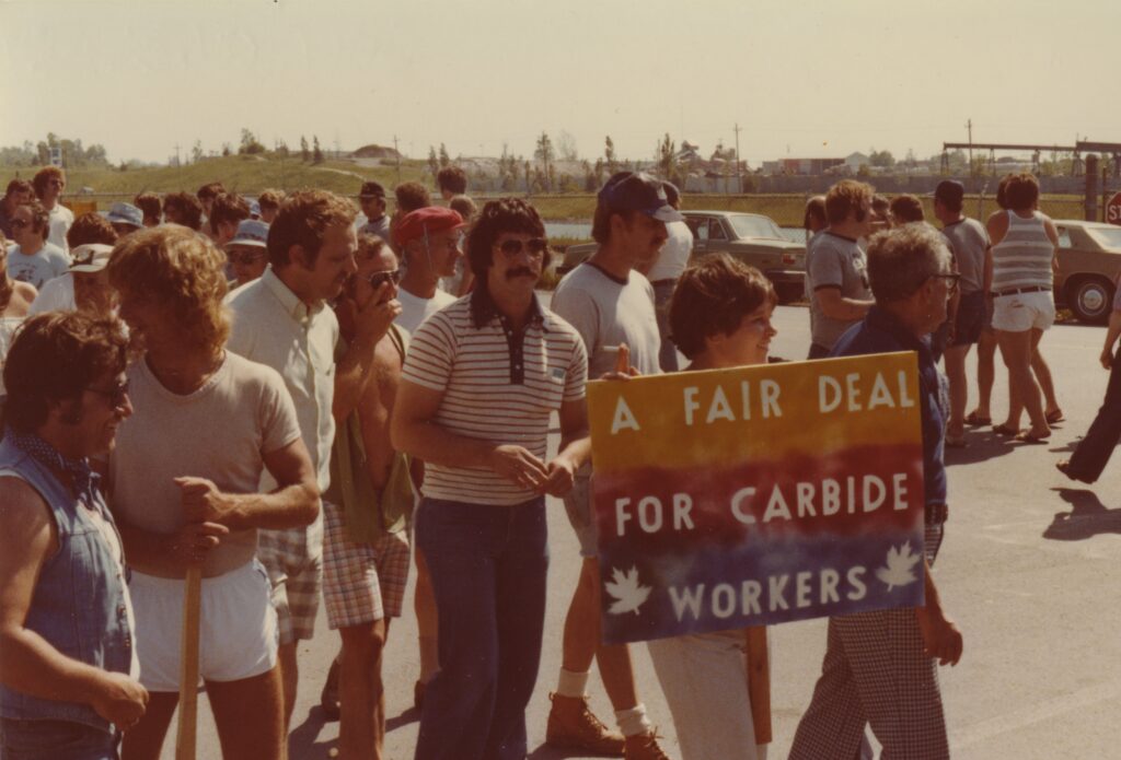 An old colour photo of a crowd of people protesting. One holds a sign that reads "a fair deal for carbide workers"