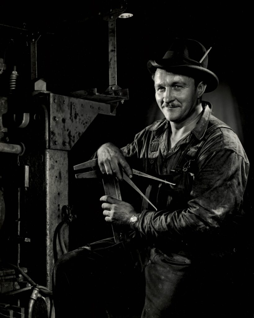 A black and white portrait of a man posing with machinery. He is wearing work clothes and a hat. 