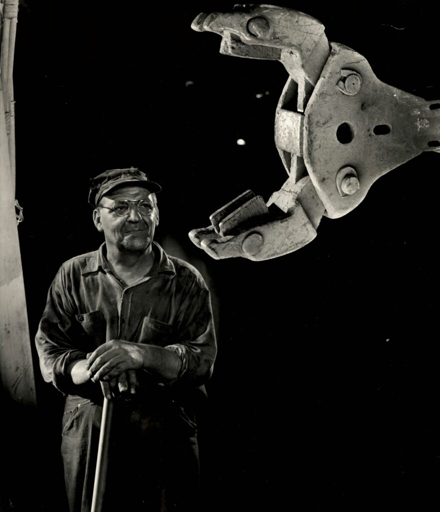 A black and white portrait of a man, posing with the claw of some sort of equipment. He is wearing a hat and glasses, and his hands rest on some sort of cane or tool.