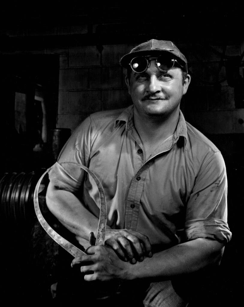 A black and white portrait of a man posing with a piece of equipment or a tool, wearing a hat and sunglasses or goggles.