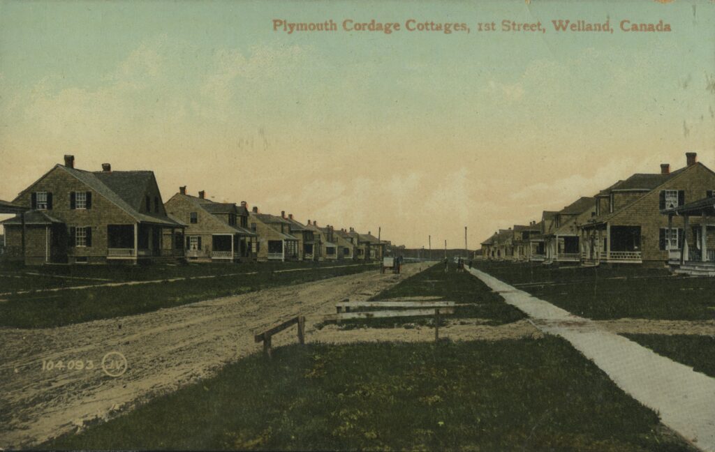 An old photo in colour with rows houses going into the distance on either side of a dirt road.