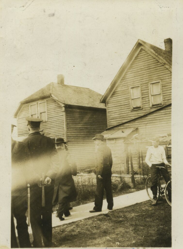 An old photo of a few people in front of a house. One looks to be a police officer, and another is on a bike.
