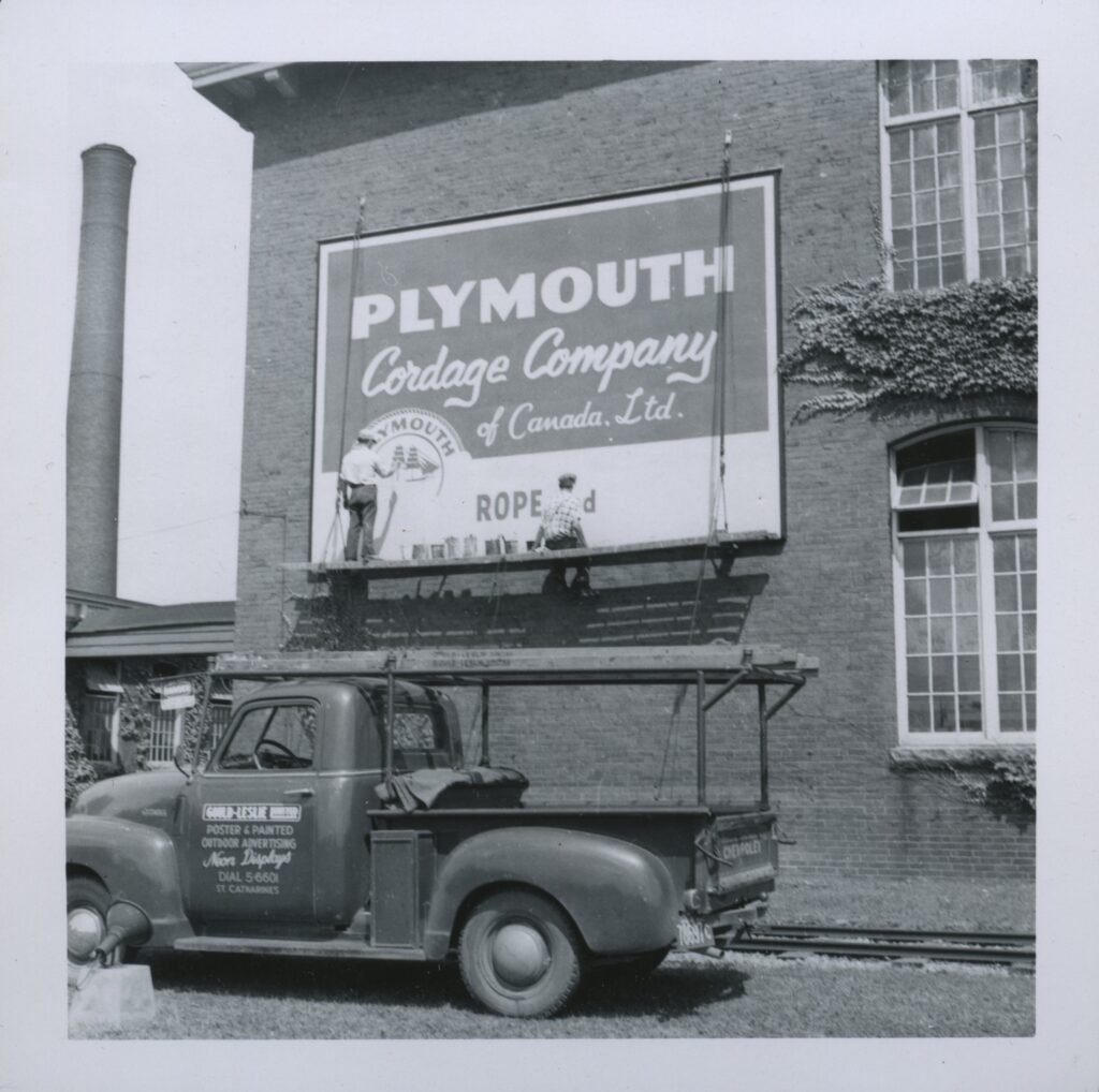 Two men work on a Plymouth cordage company billboard on the side of a building, above an old truck.