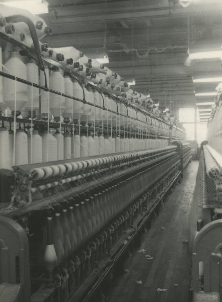 A photo of the interior of empire cotton mills, showing a couple rows of machinery and a window in the distance.